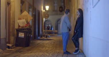 A man and a woman are standing near the wall of the night street of the old city. They talk, date. He looks around. Against the background of a trash can and a worker. CZ, Prague, Karlova, 1.10.22