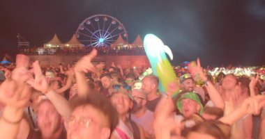 A crowd of people dance at the festival in the light. They move their hands, they have fun. In the background is a ferris wheel. Night event. CZ, Milovice, 6.8.22 Let it Roll