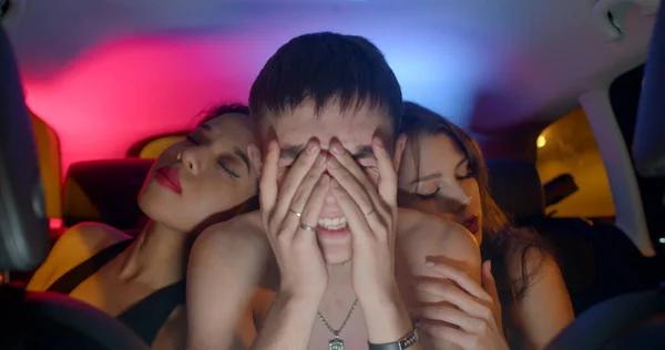 A shirtless guy sings a rap song in the car. The girls sit on the sides. They are on his shoulders. He gestures. Afro girl red lipstick. Red blue light in bokeh background.