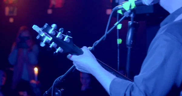 The back, the hand of the musician withstands the neck of the guitar performance. He sings a song into a microphone in blue light. Making music in public.