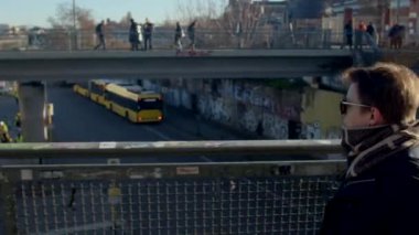 Gözlüklü bir adam şehrin köprüsü boyunca yaya köprüsünün arka planında hareket ediyor. Metal ağ çiti. DE, Berlin, Mitte, 16.1.23. 