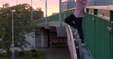 He looks down. He straightens his hair his hand guy stands on the bridge and holds handrail with his hands. Against the backdrop of the city in sunset light. CZ, Prague, Vltavska, 30.9.22 