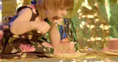 A little girl eats a cake on the table with a fork. She tilts her head to the sweet dessert, licks her fork. Golden glare background.