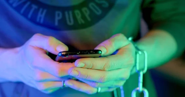 Close-up of a hand using a phone in the neon light of a nightclub. He writes a message date online registration. Chain on hand. Fingers press, scroll further.