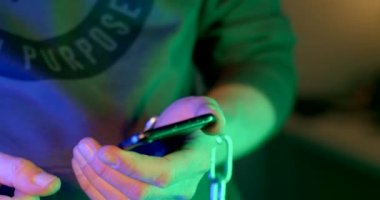 Close-up of a hand using a phone in the neon light of a nightclub. He writes a message date online registration. Chain on hand. Fingers press, scroll further.