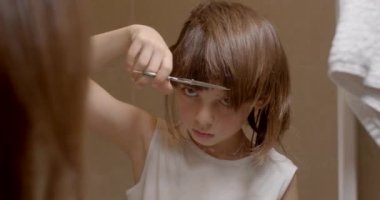 A teenage boy cuts the hair of the bangs with scissors on his face with his hands on his own. He carefully looks at the device with his eyes. Self care at home.