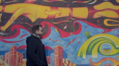 Kamera hareketindeki adam şehirdeki duvardaki resme bakıyor, kırmızı, siyah, sarı renkler. Gözlerde gözlük var. O, şehrin caddesine gider. DE, Berlin, Mitte, 16.1.23. 