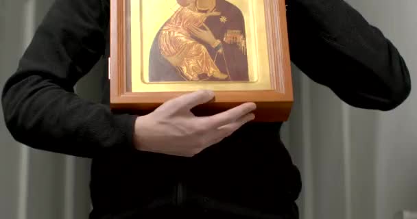 アイコンを持つ幸せな若い男の肖像祝福の母マリアと神のイエスの子 ストローブ光効果 現代のキリスト教 暗い室内 マドンナと子供の黄金の肖像画 — ストック動画