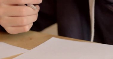 Kapüşonlu bir kişi, kalemle yazı yazarken görülüyor, çalışma alanında odak ve yaratıcılık gösteriyor.