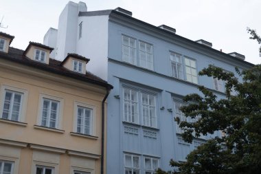 Çok sayıda penceresi olan büyük bir bina var ve önünde gölgelik ve yeşillik sağlayan güzel bir ağaç var.