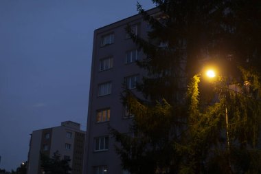 Bulanık bir resim, gece aydınlanan yüksek bir binayı ön planda belirgin bir sokak ışığı ile tasvir ediyor, eşsiz bir atmosfer yaratıyor.