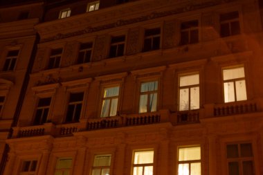 Sayısız pencereyle süslenmiş yüksek bir bina parlak, gece gökyüzüne karşı belirgin bir şekilde aydınlatılmış, güzel bir manzara yaratıyor.