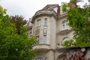 Birden fazla penceresi olan çok büyük bir bina, bölgeye gölge ve güzellik sağlayan yüksek ağaçlarla çevrilidir.