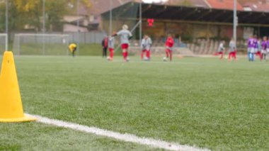 Beyaz çizgi üzerinde Sarı koni çimen sahasında arka planda futbol oyuncuları koni ve hedefleri olan bir sahada antrenman yapıyorlar, manzaralı bir arka planda.