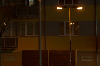 Uzun bir binanın önünde, gecenin karanlığında bir sokak lambası parıldıyor, çevreye sıcak bir parıltı saçıyor.