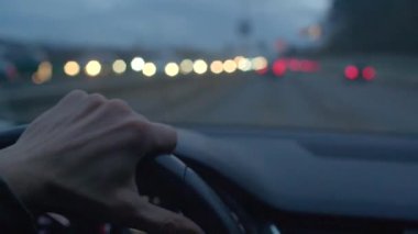 Arka planda trafik ışıkları olan bir aracın içindeki direksiyonu sıkıca tutan bir el.