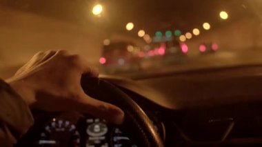 Gece bir tünelde, bir el direksiyonu kavrıyor trafik akarken, ilerideki şehir yolculuğunu işaret ediyor.