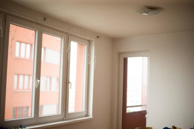 Işıl ışıl aydınlatılmış bir iç mekan büyük pencereler ve çarpıcı bir kırmızı binaya açılan bir kapı sergiliyor.