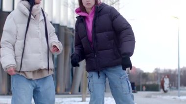 Kış kıyafetleri içinde iki neşeli genç, modern bir binanın yanında poz veriyor, günün tadını çıkarıyorlar.
