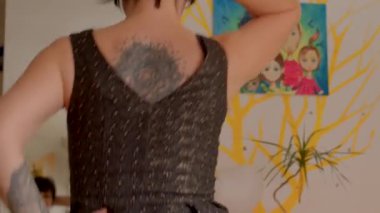 Siyah saçlı dövmeli bir kadın, zarif giyinmiş, arka planda büyüleyici bir sanat eserinin ortasında oturuyor.