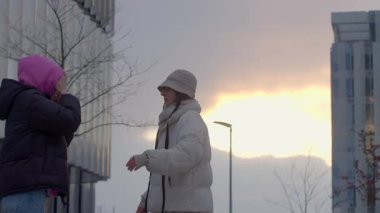 İki insan neşeli bir şekilde dışarıda poz veriyor, canlı bir şehir ortamında çarpıcı bir günbatımı tarafından çerçevelenmiş.