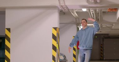 Bir adam, arka planda sarı uyarı bandı olan bir otoparkta rastgele birini selamlıyor.