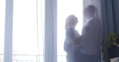 Bir çift, arka plandaki pencerenin yanında sıcak bir kucaklaşmayı paylaşıyor. Yumuşak bir arka plan onların samimiyetini arttırıyor.