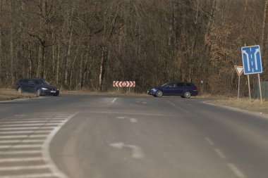 Aydınlık bir günde, mavi bir araba durmadan ağaçlarla dolu yemyeşil bir orman boyunca uzanan dolambaçlı bir yolda ilerliyor.