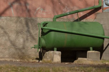 Yeşil silindirik bir tank dayanıklı tuğla bir duvarın yanında duruyor ve etrafındaki renk ve dokularda ilginç bir kontrastlık oluşturuyor.