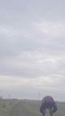 Bir birey engin bir kırsal alanda çömelmiş, arkasında dramatik bulutlu bir gökyüzü var.