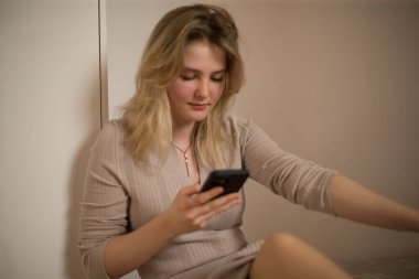 Sıradan ve basit bir iç mekan ortamında, genç bir kadın akıllı telefon cihazına derinden odaklanır.