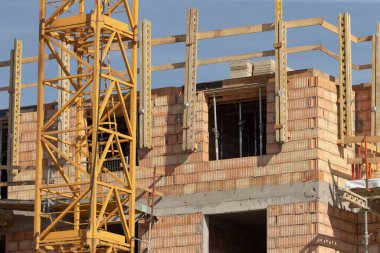 Şu anda büyük bir tuğla bina inşa ediliyor ve ön planda parlak sarı bir vinç yer alıyor.