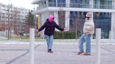 Karlı bir şehir ortamında, kışın giyinmiş iki kişi binaların arasında dikiliyor.