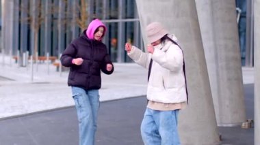 Kapüşonlu ceket giyen iki kişi açık havada, şehir mimarisinin arka planına karşı.