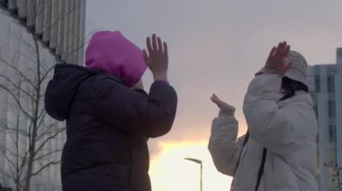 İki kadın dışarıda şık kış kıyafetleri içinde güzel bir şehrin günbatımı arkaplanıyla sıcak bir şekilde etkileşime girer.
