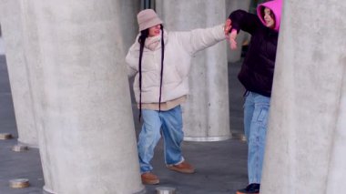 Kentsel bir ortamda, iki çocuk beton sütunların arkasında eğlenceli bir etkileşim içindeler.