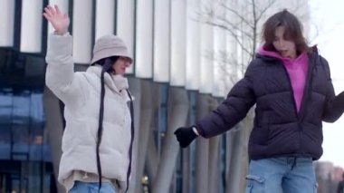Sıcak kış kıyafetleri giymiş iki kişi canlı bir şehir ortamında eğlenceli bir şekilde etkileşime geçiyorlar.