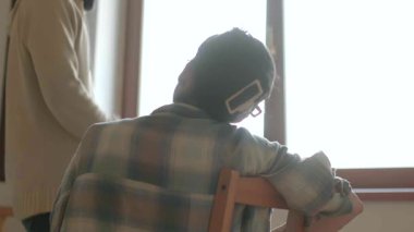 Pencere arkası olan bir odada, iki birey bir sohbete dahil oluyor, anlamlı bir şekilde etkileşime geçiyorlar.