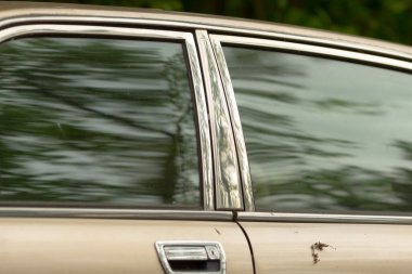 Bu görüntü, arka planda belirgin yeşil ağaçlar bulunan ve sakin bir atmosfer yaratan bir araba penceresinin detaylı yakın çekimini içeriyor.