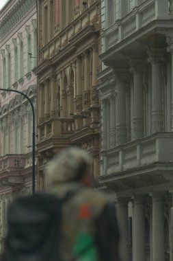 Bir adam hareketli bir caddede boş boş yürüyor, yol boyunca uzanan çeşitli binaların önünden manzarayı seyrediyor.