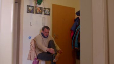 Sessiz bir koridorda, paltolu bir adam düşünmek için durur, ayakkabılarını bir tabureye bağlar. Bu huzurlu an, onun durgunluktan harekete geçişini vurguluyor.