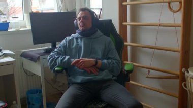 Kulaklık takan odaklanmış bir adam bilgisayarın önünde sandalyeye oturur. Modern bir çalışma alanında gayret ve adanmışlık gösteriyor.