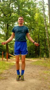 Mavi elbiseli bir adam parkta güçlü bir ip atlama egzersizi yapar. Sonra da telefonunu sakin, ağaçlık bir alanda kontrol eder.