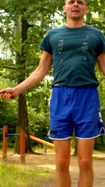 Video, mavi tişörtlü ve şortlu bir adamın bir parkta atladığını ve çeviklik gösterdiğini gösteriyor. Yakınlarda, yeşil tişörtlü bir adam izliyor. Açık hava sporunu somutlaştırıyor.