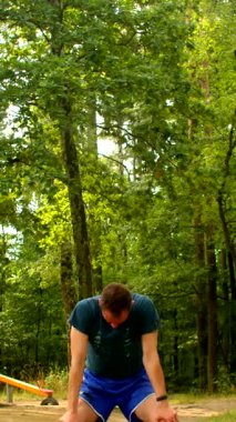 Yeşil tişörtlü ve mavi şortlu bir adam gür bir ormanda diz çöker, egzersiz yapmak için eğilir, sonra da dingin ağaçların arasında şınav çeker.