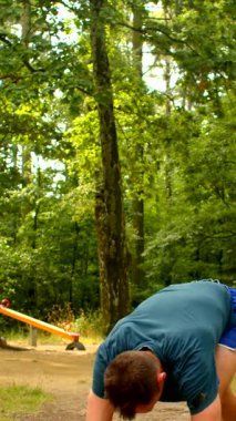 Mavi tişörtlü ve şortlu bir adam canlı bir parkta spor yapıyor. İp atlama ve şınav çekerek gücünü ve bağlılığını gösteriyor.