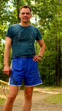 Videoda mavi şortlu ve yeşil gömlekli bir adam manzaralı toprak bir yolda atlama ipi ve tenis raketiyle atletizmini sergiliyor.