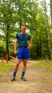 Mavi şortlu ve gömlekli bir adam yemyeşil ormandaki toprak bir yolda, elinde ip ve tenis raketi tutarak, açık hava fitnesinin keyfini çıkarıyor.