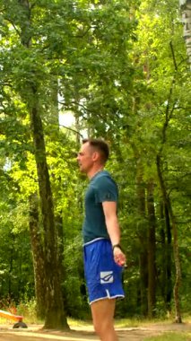Mavi şortlu bir adam, sakin bir ormanda egzersiz yapıyor, esniyor, eğiliyor ve ip atlıyor, yemyeşil halatla açık hava fitness temasını geliştiriyor.