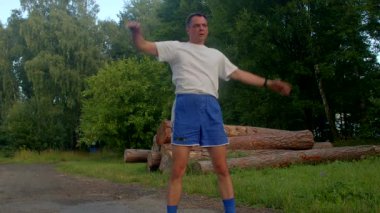 Beyaz tişörtlü ve mavi şortlu bir adam, sakin bir parkta kollarını ve bacaklarını uzatarak, doğadaki uygunluğunu ve sağlığını ön plana çıkarıyor.